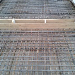 Siatka zbrojeniowa na budowie, z drewnianym szalunkiem w górnej części kadru. Widok z góry, ukazujący regularny układ prętów.