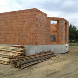 Budowa domu w stanie surowym otwartym: ściany z cegły ceramicznej, w tle drzewa i niebo. Na pierwszym planie stosy drewnianych bali.