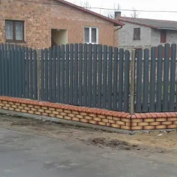 Szary, metalowy płot lamelowy z podmurówką z cegły klinkierowej w Konopnicy, widok z poziomu ulicy, z domami w tle.