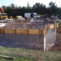 Budowa fundamentów: Ściany fundamentowe z bloczków betonowych, szalunek z desek, zbrojenie siatką stalową, w tle betoniarka i pracownicy.