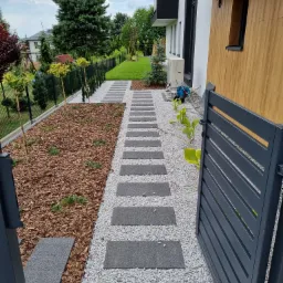 Nowoczesny ogród w Katowicach z ścieżką z płyt betonowych, wysypaną białym żwirem, obrzeżem z kory i młodymi drzewkami wzdłuż ogrodzenia.