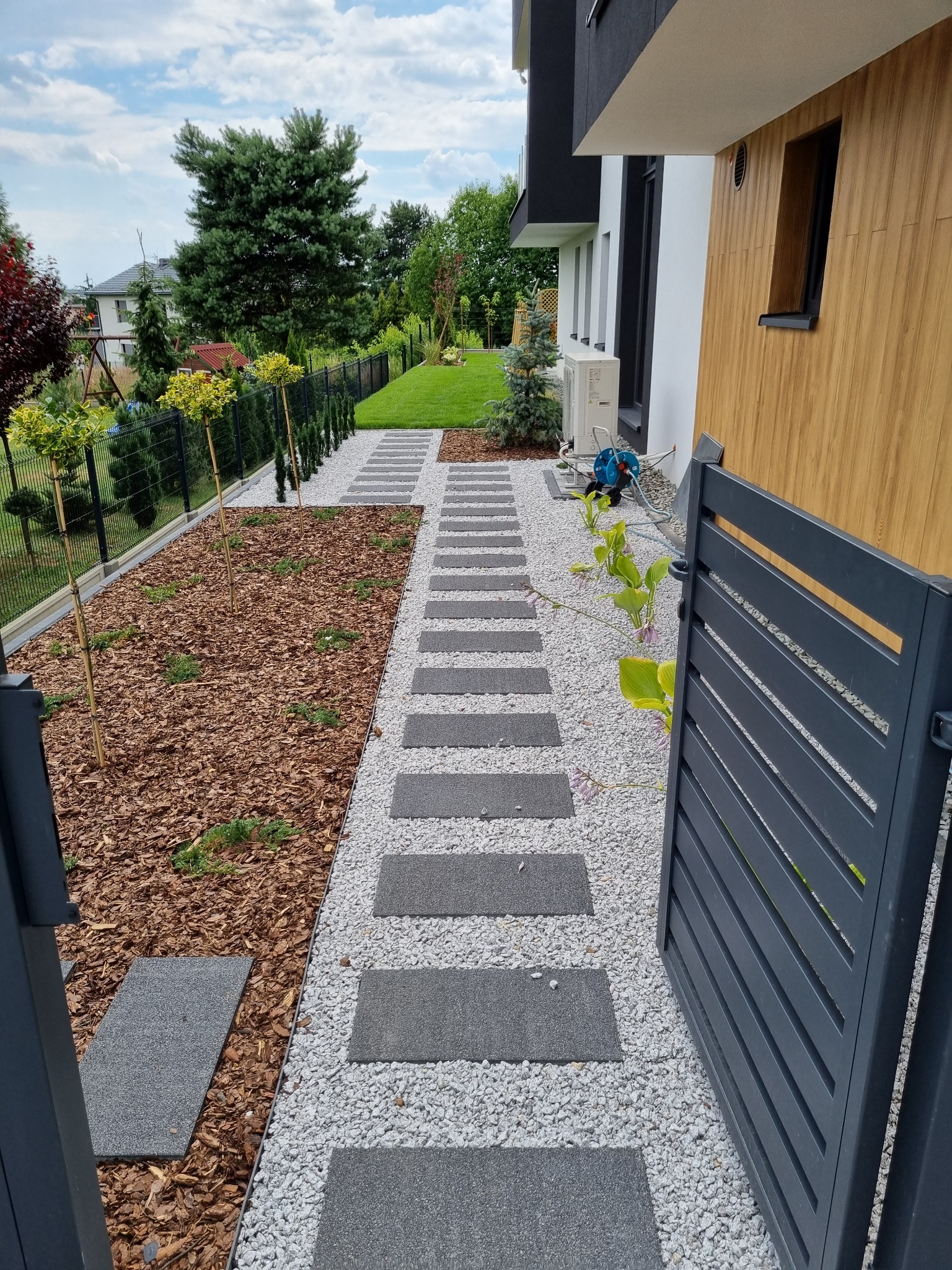 Nowoczesny ogród w Katowicach z ścieżką z płyt betonowych, wysypaną białym żwirem, obrzeżem z kory i młodymi drzewkami wzdłuż ogrodzenia.