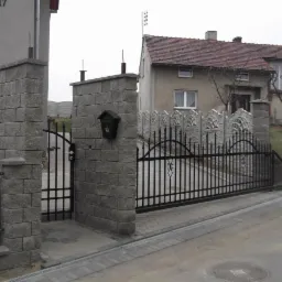 Nowoczesna brama wjazdowa z czarnymi, kutymi elementami i szarymi, kamiennymi słupkami. Kostka brukowa w odcieniach szarości prowadzi do posesji.