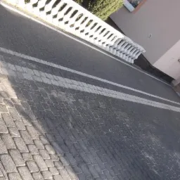 Szara kostka brukowa ułożona w regularny wzór, z białymi pasami akcentującymi, tworząca podjazd do budynku z balustradą. Widoczny numer domu.