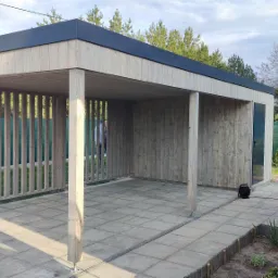 Nowoczesna altana z jasnego drewna z elementami ażurowymi, z ciemnym płaskim dachem, na wyłożonym kostką tarasie, z widocznym kablem elektrycznym i narzędziami w tle.