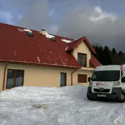 Dom jednorodzinny z nowym, czerwonym dachem z blachodachówki, częściowo pokryty śniegiem, z oknami dachowymi. Na pierwszym planie biały bus firmy z otwartymi drzwiami w zimowej scenerii.