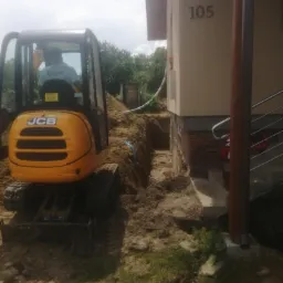Żółta mini koparka JCB wykonuje wykop pod fundament budynku z numerem 105, widoczne odsłonięte fundamenty i fragment instalacji.