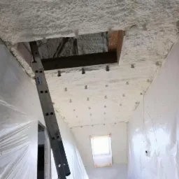 Poddasze zabezpieczone folią, pokryte białą pianą izolacyjną, widoczny fragment drabiny i otwór w stropie z belką konstrukcyjną.