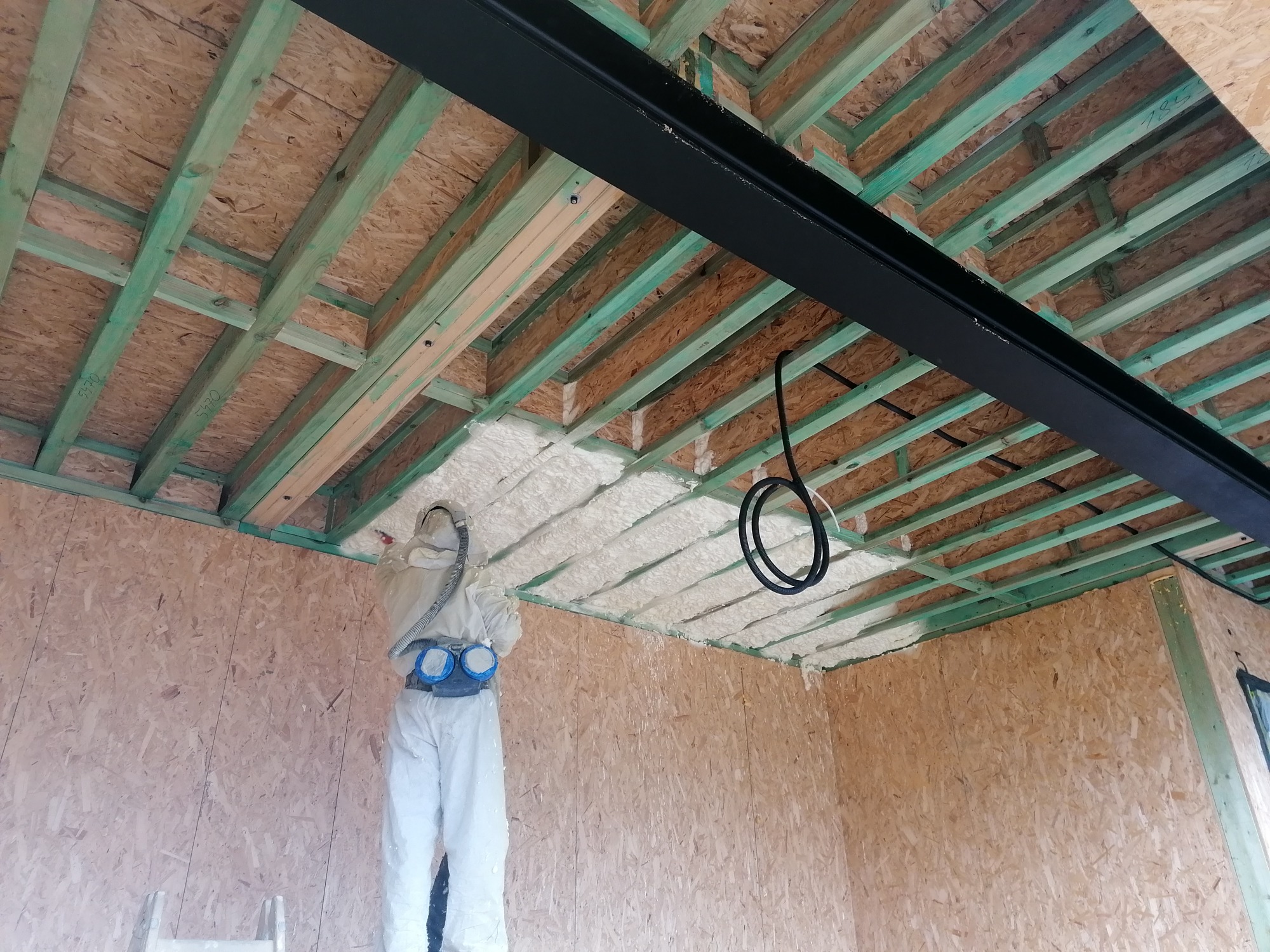 Pracownik w kombinezonie ochronnym natryskuje białą pianę izolacyjną na drewniany strop z zielonymi belkami wewnątrz budynku, widoczna czarna belka konstrukcyjna i przewody elektryczne.