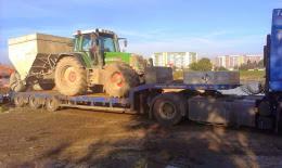 Zielony traktor Fendt z doczepionym zbiornikiem na platformie niskopodwoziowej, widok z boku, w tle budynki mieszkalne.