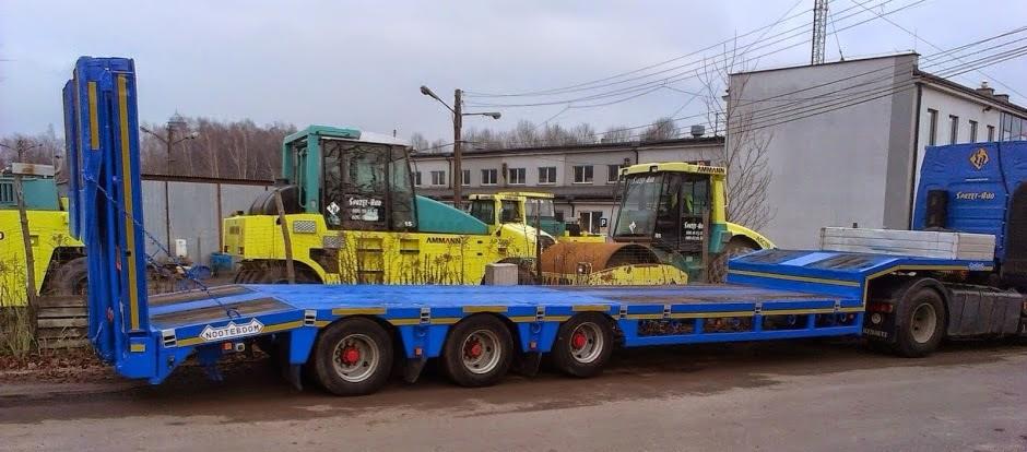 Niebieska naczepa niskopodwoziowa marki Nooteboom na tle zaparkowanych walców drogowych Ammann i budynku przemysłowego.