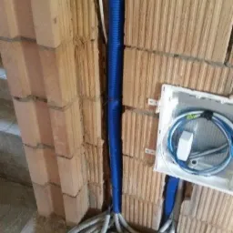 Niebieska rura karbowana pionowo zamocowana do ściany z cegieł, biała puszka instalacyjna z niebieskimi przewodami, wiązka szarych rur karbowanych na podłodze.