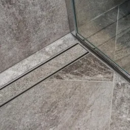 Szczeg&oacute;łowe ujęcie narożnika kabiny prysznicowej z szarymi, matowymi płytkami imitującymi kamień, z widocznym odpływem liniowym i uszczelnieniem szkła kabiny.