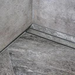 Szczegół ułożenia szarych, matowych płytek w rogu łazienki z odpływem liniowym, widoczne fugi i tekstura płytek.