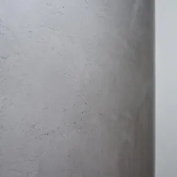 Szara, teksturowana ściana z widocznymi nierównościami i delikatnymi smugami, przylegająca do białej ramy okiennej, z zielenią widoczną za oknem.