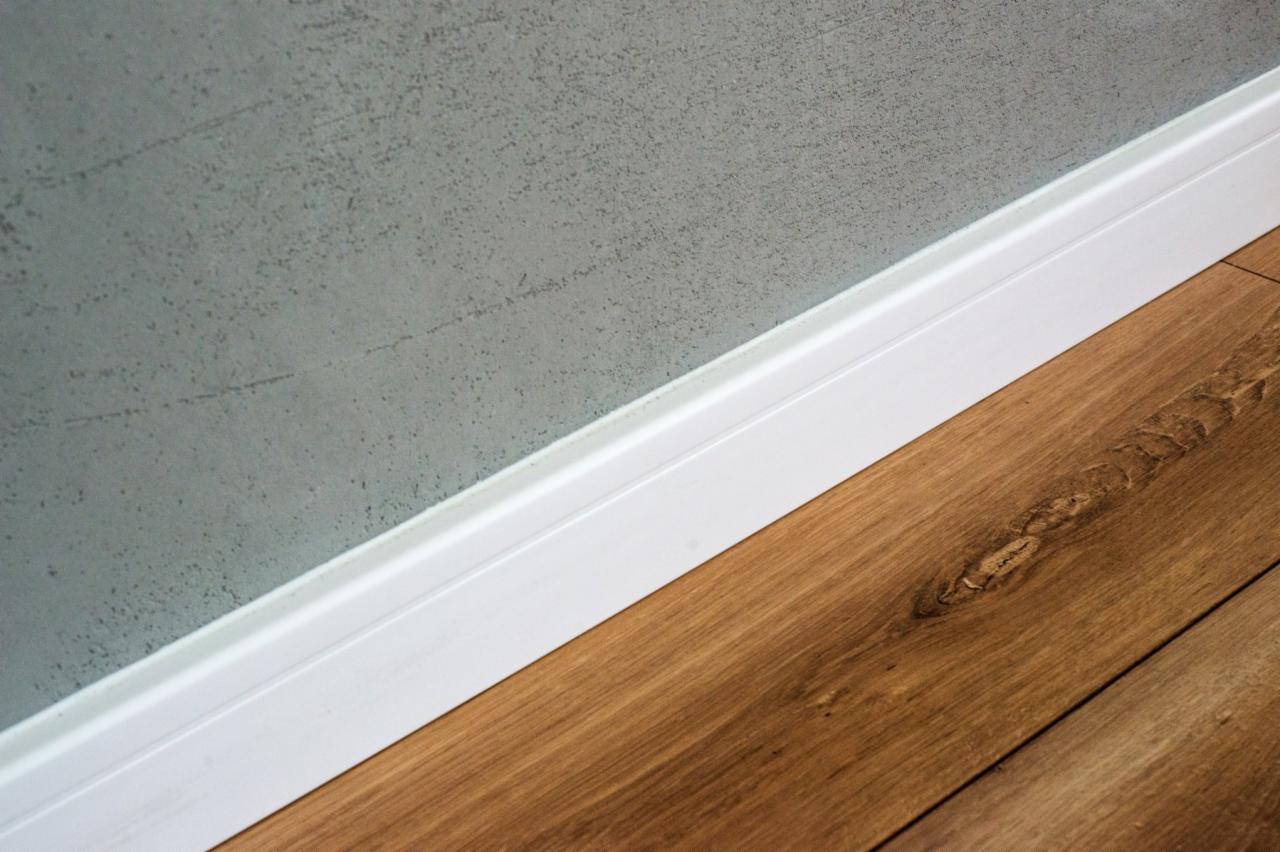 Szary tynk dekoracyjny na ścianie z białą listwą przypodłogową przy parkiecie z wyraźnym usłojeniem drewna.