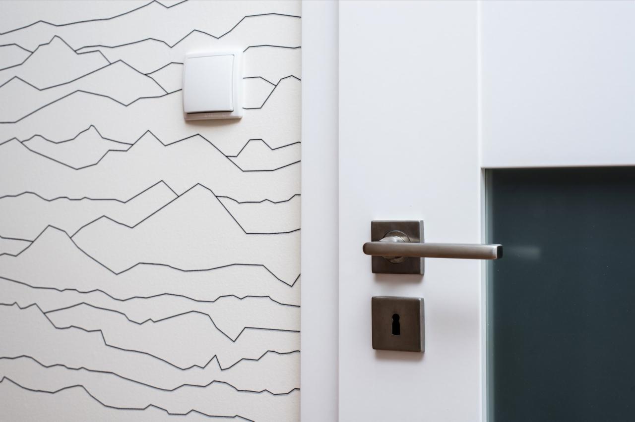 Biała ściana z wzorem gór w stylu line art, biały włącznik światła i fragment białych drzwi z nowoczesną, kwadratową klamką w kolorze grafitowym.