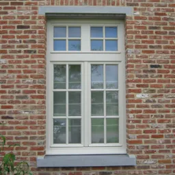 Białe, wielokwaterowe okno wkomponowane w czerwoną, ceglaną ścianę z szarym kamiennym parapetem. Widok z zewnątrz, drzewa odbijają się w szybach.