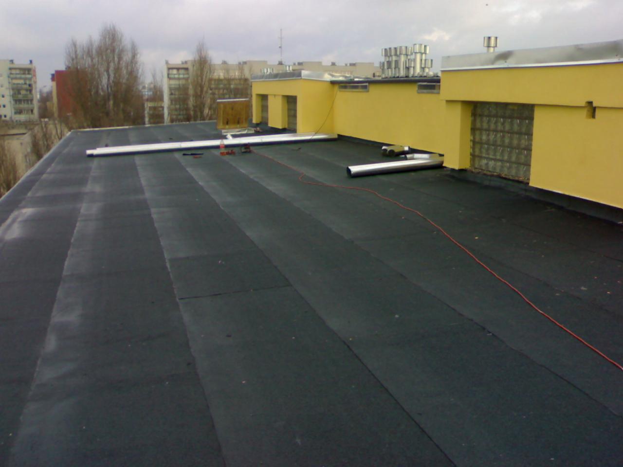 Czarny, płaski dach pokryty papą termozgrzewalną z widocznymi narzędziami i elementami blacharskimi na tle miejskiego krajobrazu.