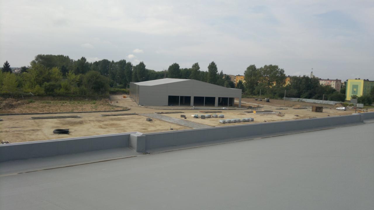 Nowoczesny, szary budynek magazynowy w trakcie budowy, widziany z góry. W tle zieleń i zabudowa miejska. Widoczny fragment płaskiego dachu w pierwszym planie.