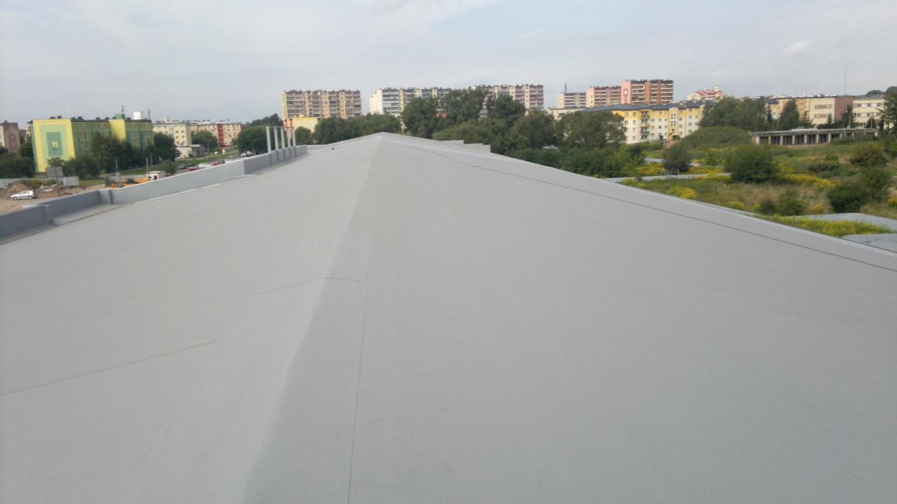 Szary, płaski dach budynku z widocznymi detalami łączeń i obróbek blacharskich na tle zabudowy miejskiej i zieleni. Ujęcie z góry.