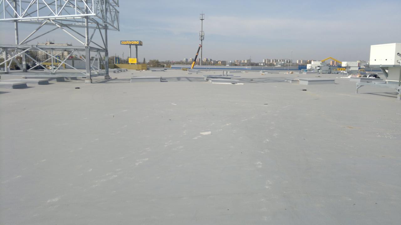 Szary, płaski dach budynku komercyjnego z elementami konstrukcyjnymi i wentylacyjnymi. W tle widoczny dźwig i zabudowa miejska.