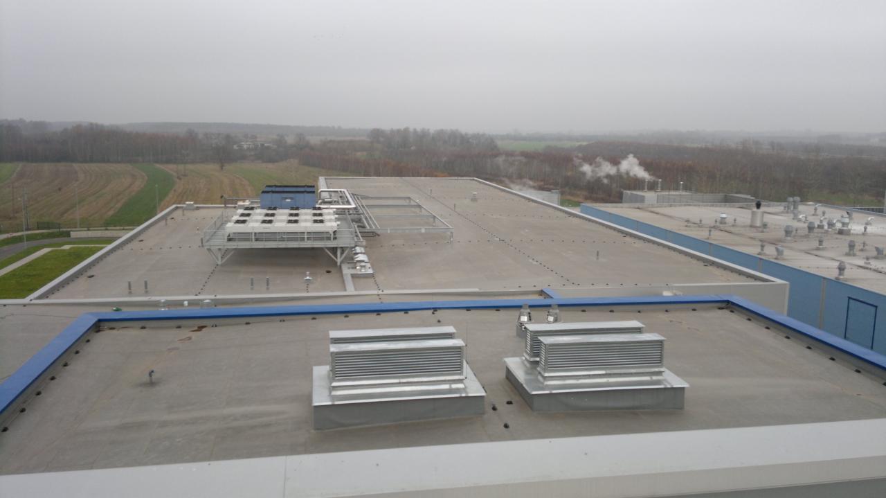 Widok z góry na płaski dach budynku przemysłowego z instalacjami wentylacyjnymi i technicznymi. W tle pola i las pod mglistym niebem.