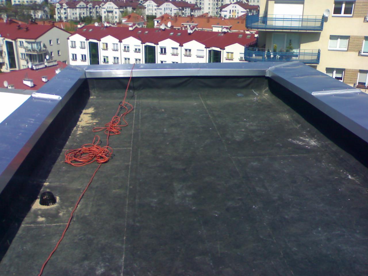 Płaski dach pokryty czarną papą, zabezpieczony metalową obróbką. Na dachu leży zwój czerwonego sznurka. W tle zabudowa miejska z czerwonymi dachami.