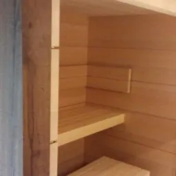 Wnętrze sauny z jasnego drewna, z dwoma poziomami ławek. Widoczne detale konstrukcyjne i wykończeniowe, naturalne oświetlenie, surowe drewno.