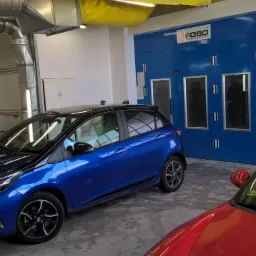 Niebieski samochód Toyota Yaris w warsztacie przed kabiną lakierniczą FOBO, obok zaparkowane czarne auto i czerwone auto sportowe.