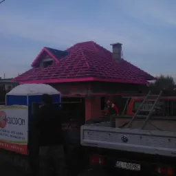 Rozpoczęta budowa dachu z widoczną konstrukcją drewnianą w kolorze różowym, częściowo pokryta dachówką, na tle budynków mieszkalnych i samochodu dostawczego z drabiną na pace.