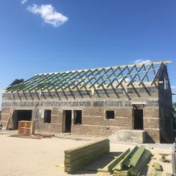 Budowa domu w stanie surowym otwartym z bloczków, widoczne drewniane elementy konstrukcji dachu, stos belek i pustaków na placu budowy.