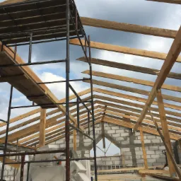 Wznoszenie konstrukcji dachu z drewnianych belek na budynku z białych bloczków, widoczne rusztowanie i pochmurne niebo.