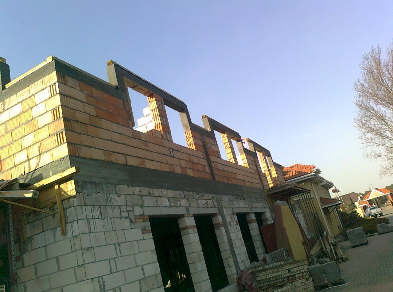 Wznoszenie ścian domu murowanego z bloczków ceramicznych, widoczne otwory okienne z nadprożami, na pierwszym planie białe bloczki silikatowe, w tle inne budynki i błękitne niebo.