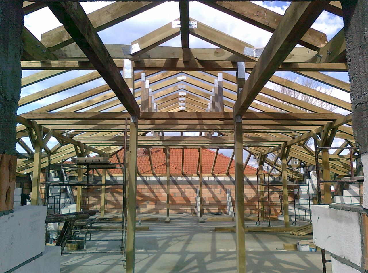 Wnętrze budynku w trakcie budowy z widoczną drewnianą konstrukcją dachu, metalowymi łącznikami, częściowo wymurowanymi ścianami oraz rusztowaniami w tle.