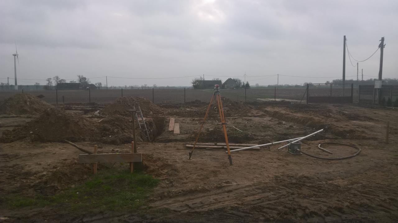 Wykop pod fundamenty z widocznym niwelatorem laserowym na trójnogu, rurami i stertami ziemi na budowie w pochmurny dzień. W tle dom i turbiny wiatrowe.