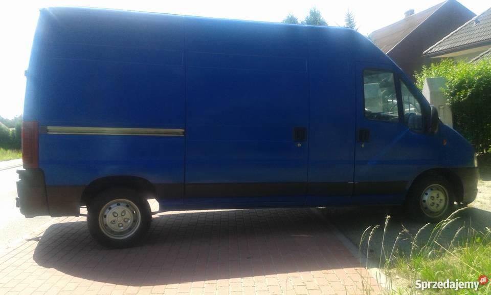 Niebieski samochód dostawczy zaparkowany na chodniku z kostki, widok z boku, z widocznym logo 'S' w prawym dolnym rogu zdjęcia.