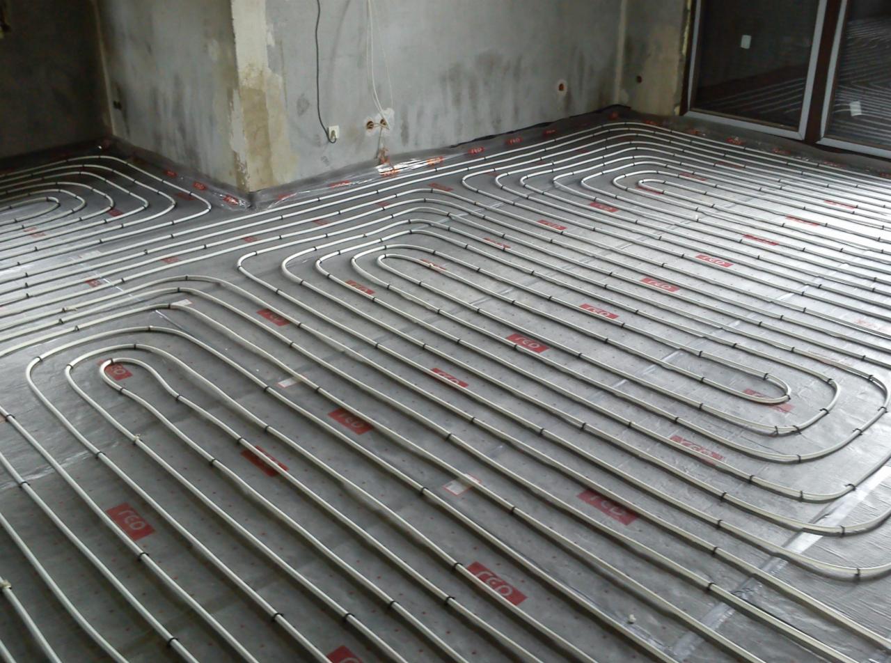 Układanie rur ogrzewania podłogowego na folii izolacyjnej w surowym wnętrzu. Widoczne betonowe ściany i duże okno balkonowe. Etap przed wylewką.