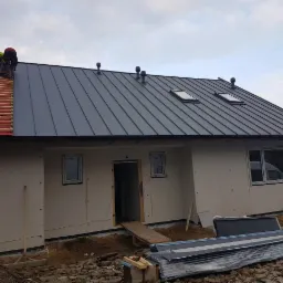 Montaż szarej blachy na dachu domu jednorodzinnego, widoczni dekarze w trakcie pracy, część dachu pokryta czerwoną konstrukcją z łat, w tle pochmurne niebo.