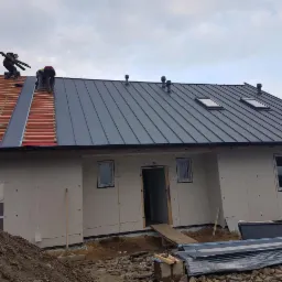 Montaż szarego pokrycia na dachu domu jednorodzinnego, widoczni dekarze pracujący na częściowo ukończonej konstrukcji, z oknami dachowymi i kominami wentylacyjnymi.