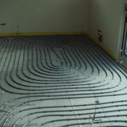 Instalatorhydraulik - Szare rury ogrzewania podłogowego ułożone spiralnie na szarej macie w jasnym pomieszczeniu z dużym oknem. Żółta taśma zabezpiecza ściany.