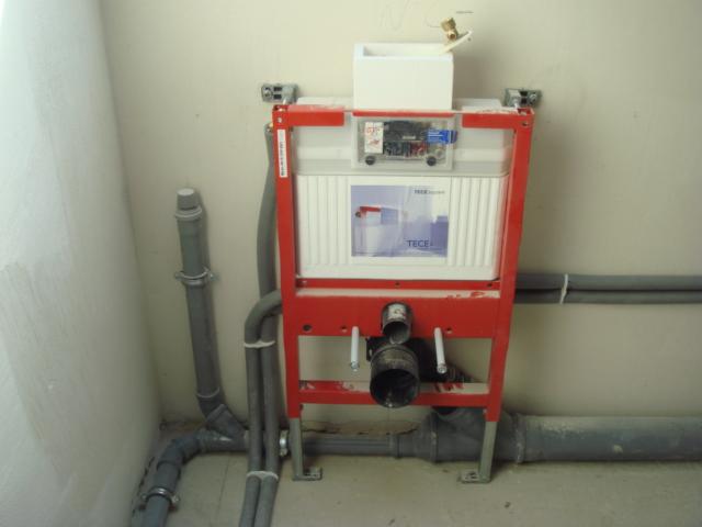 Czerwona rama stelaża podtynkowego TECE z szarymi rurami kanalizacyjnymi na tle surowej ściany, widoczne przyłącza i elementy instalacji.