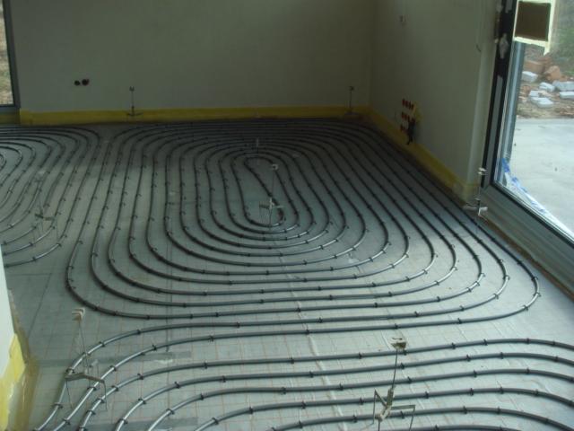 Szare rury ogrzewania podłogowego ułożone spiralnie na szarej macie w jasnym pomieszczeniu z dużym oknem. Żółta taśma zabezpiecza ściany.