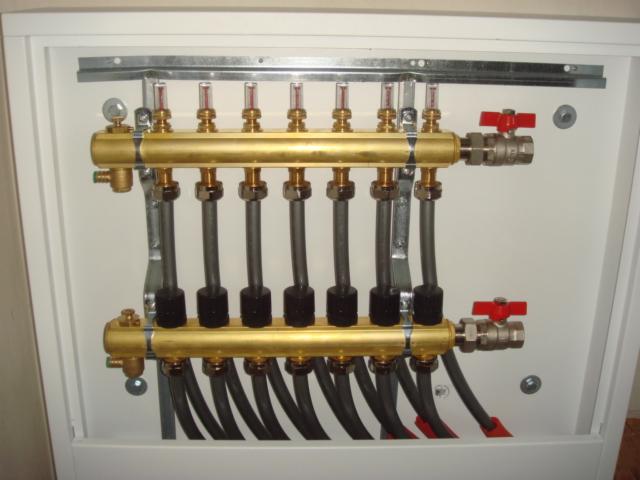 Rozdzielacz ogrzewania podłogowego w szafce, z widocznymi zaworami, rurami i elementami regulacyjnymi. Instalacja w białej szafce, widok z przodu.