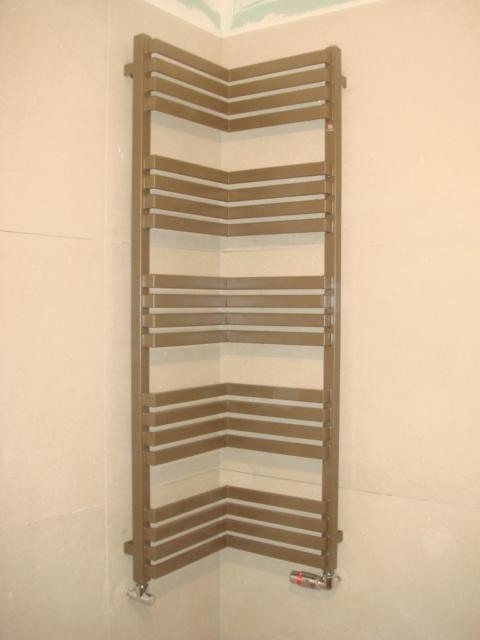 Kątowy, brązowy grzejnik łazienkowy zamontowany na jasnej, wykafelkowanej ścianie. Widoczne zawory. Nietypowy kształt dopasowany do rogu pomieszczenia.