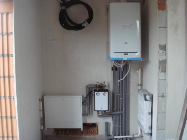 Biały piec gazowy zamontowany na ścianie w surowym wnętrzu. Widoczne rury, przewody i elementy instalacji grzewczej. Częściowo otynkowane ściany i surowy mur.