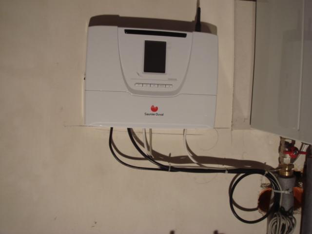 Biały regulator temperatury Saunier Duval zamontowany na ścianie, z widocznymi kablami i fragmentem instalacji hydraulicznej w tle. Ujęcie z bliska.