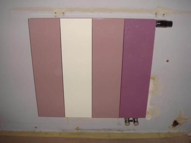 Kolorowy grzejnik panelowy na ścianie, z pionowymi pasami w odcieniach różu i beżu. Widoczne rury doprowadzające. Ściana wymaga odświeżenia.