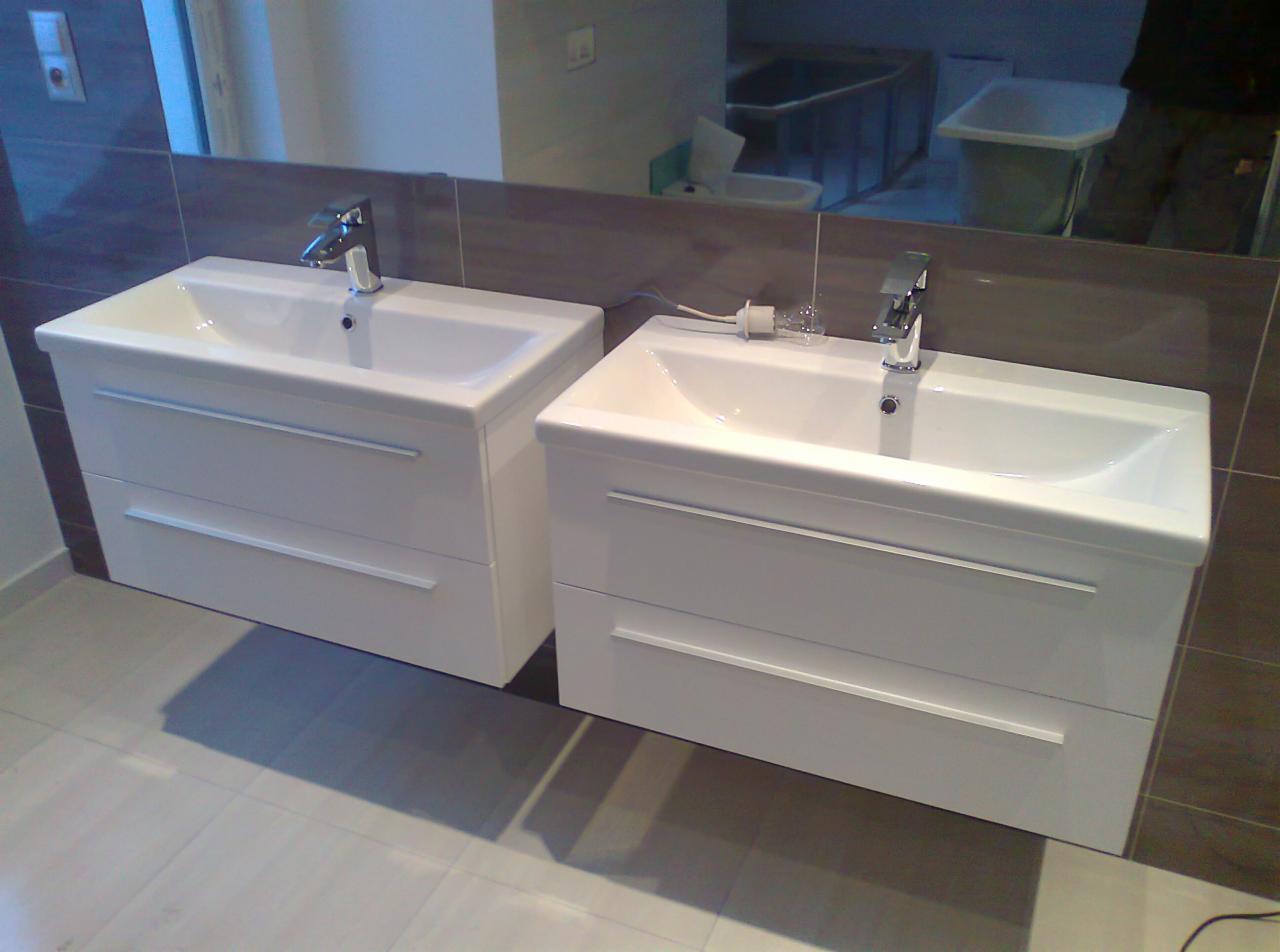 Dwie białe umywalki z szafkami i chromowaną armaturą na tle brązowych płytek i lustra. Widoczny fragment podłogi z jasnych płytek.