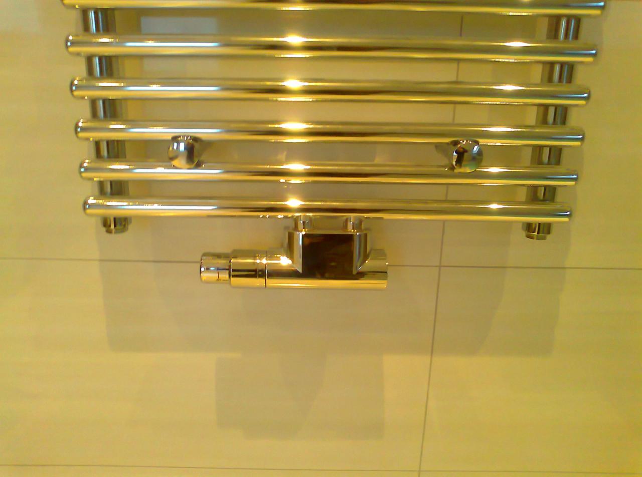 Błyszczący, złoty grzejnik łazienkowy drabinkowy z termostatem na tle płytek. Widoczne chromowane elementy i odbicia światła.